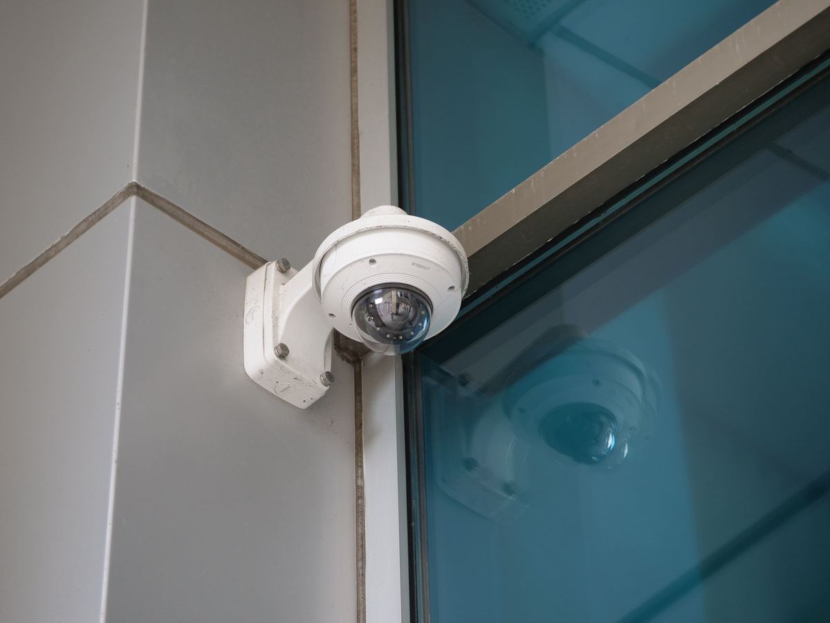 Een koepelcamera aan een gevel, met de gevel erachter in het blauwgroen.