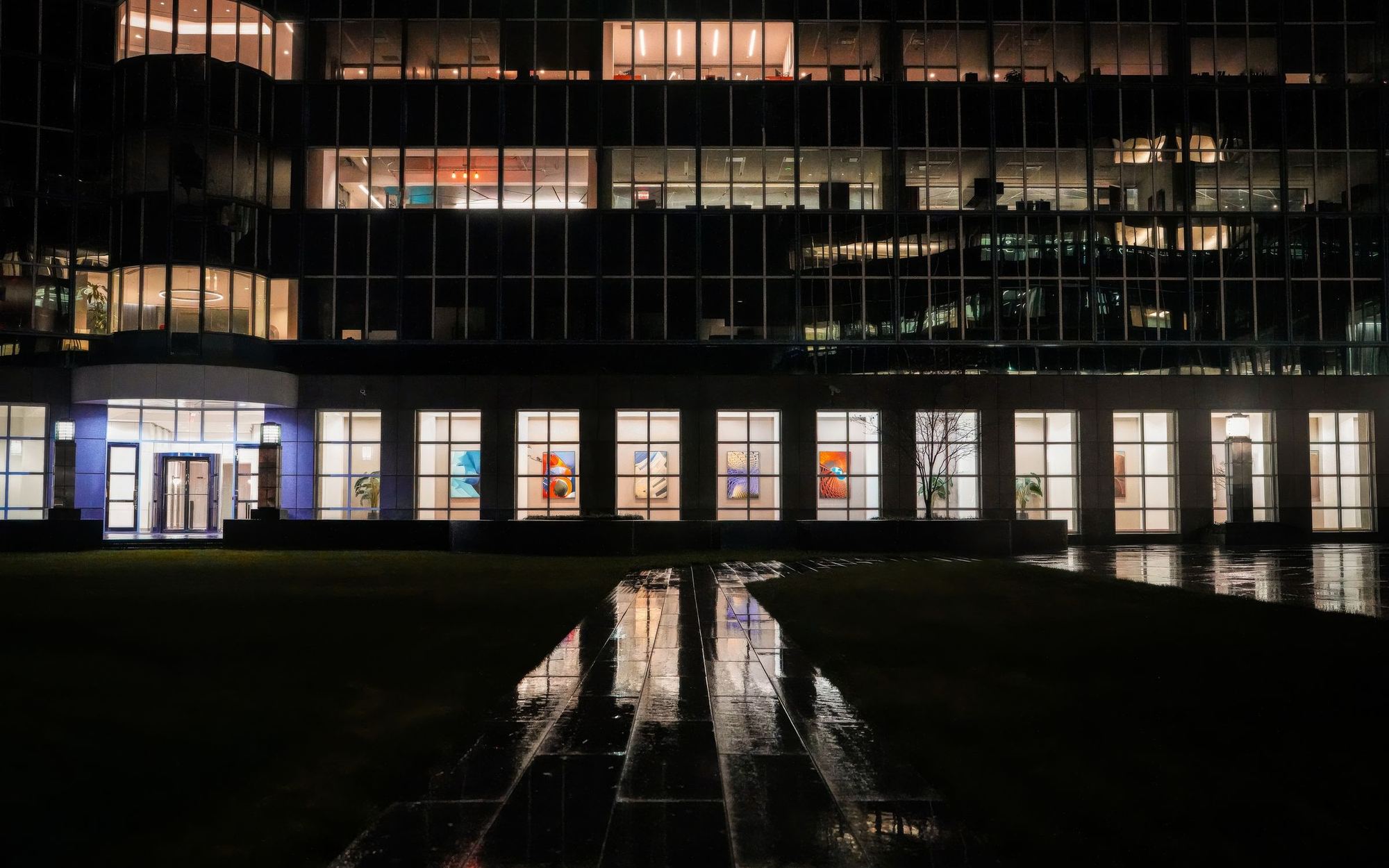 Een verlicht kantoorpand bij nacht, met de lichten weerspiegeld in het natte plein ervoor.