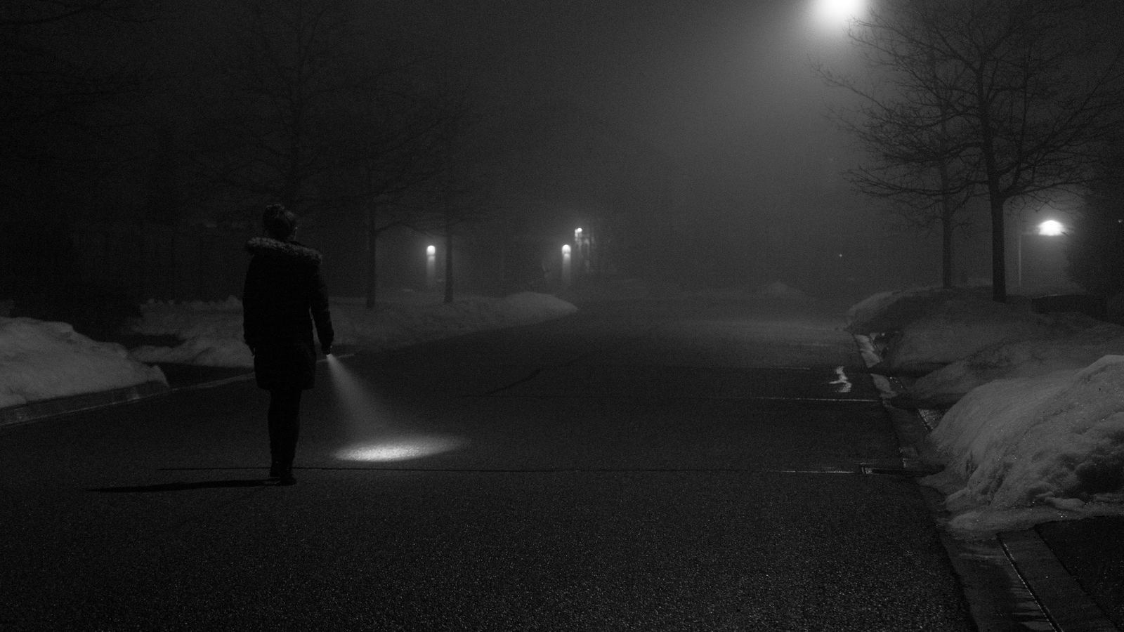 Een gestalte met een zaklamp loopt in de mist over een donkere weg.